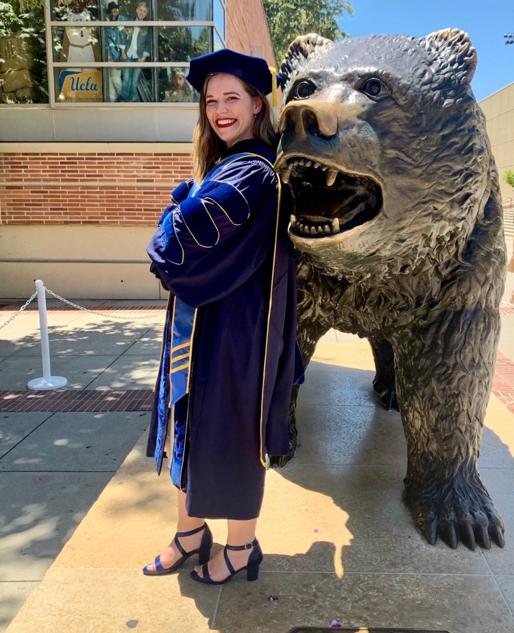 Above image shows Annie Wofford in navy and gold doctoral regalia, leaning on a statue of a bear on the UCLA campus
