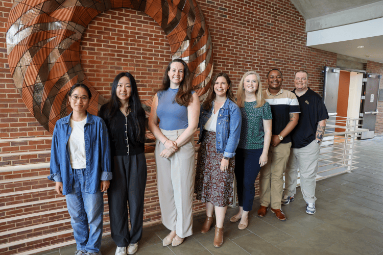 An image of the GEMS research team, including seven people standing in front of a decorative brick wall and railing in an academic building. The people are standing in a horizontal line next two each other, with two Asian women, three white women, one Black man, and one trans white researcher (from left to right).